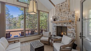 A cozy room with a stone fireplace, wicker furniture, and large windows overlooking a scenic outdoor area. - Hemlock Mist: Modern Lakeside Home w/  Screened Porch, Kayak (Highlands)