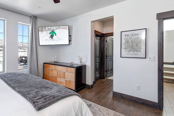 Une chambre à coucher moderne avec un grand lit, un téléviseur fixé au mur, une commode en bois et une œuvre d’art encadrée.