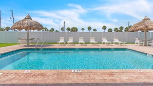 A swimming pool with lounge chairs and thatched umbrellas, surrounded by a white fence and palm trees.