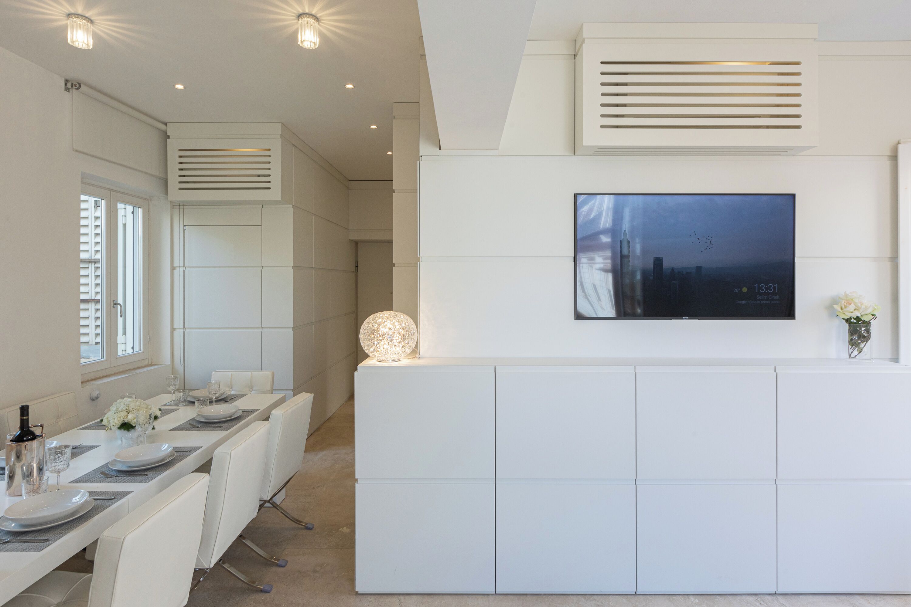 Un espace de salle à manger moderne avec une longue table dressée pour six personnes, des chaises blanches, un téléviseur à écran plat fixé au mur et une lampe sphérique décorative posée sur la table.