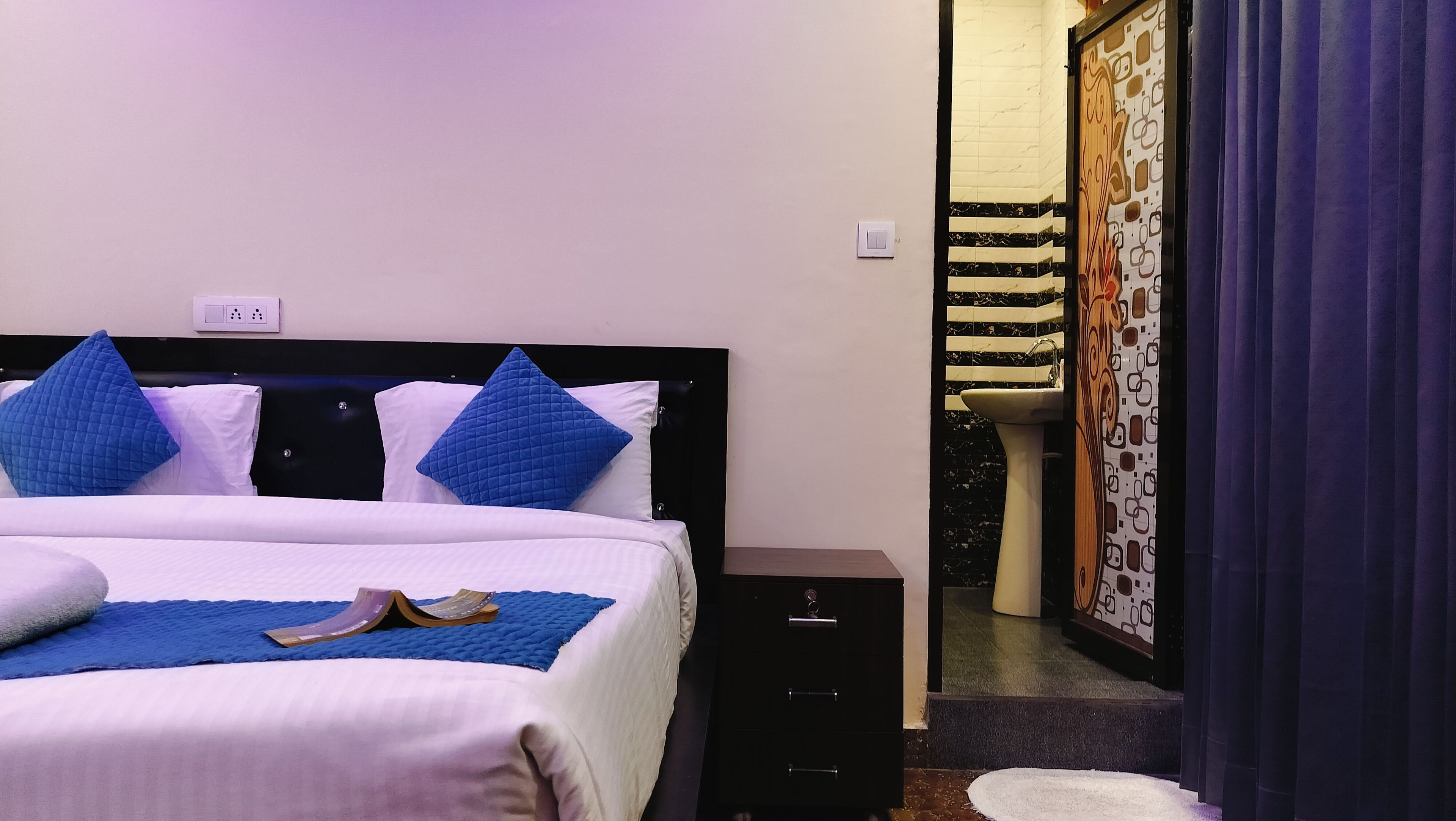 A hotel room with a bed, a nightstand, a bathroom with a sink and mirror, and a door with a curtain.