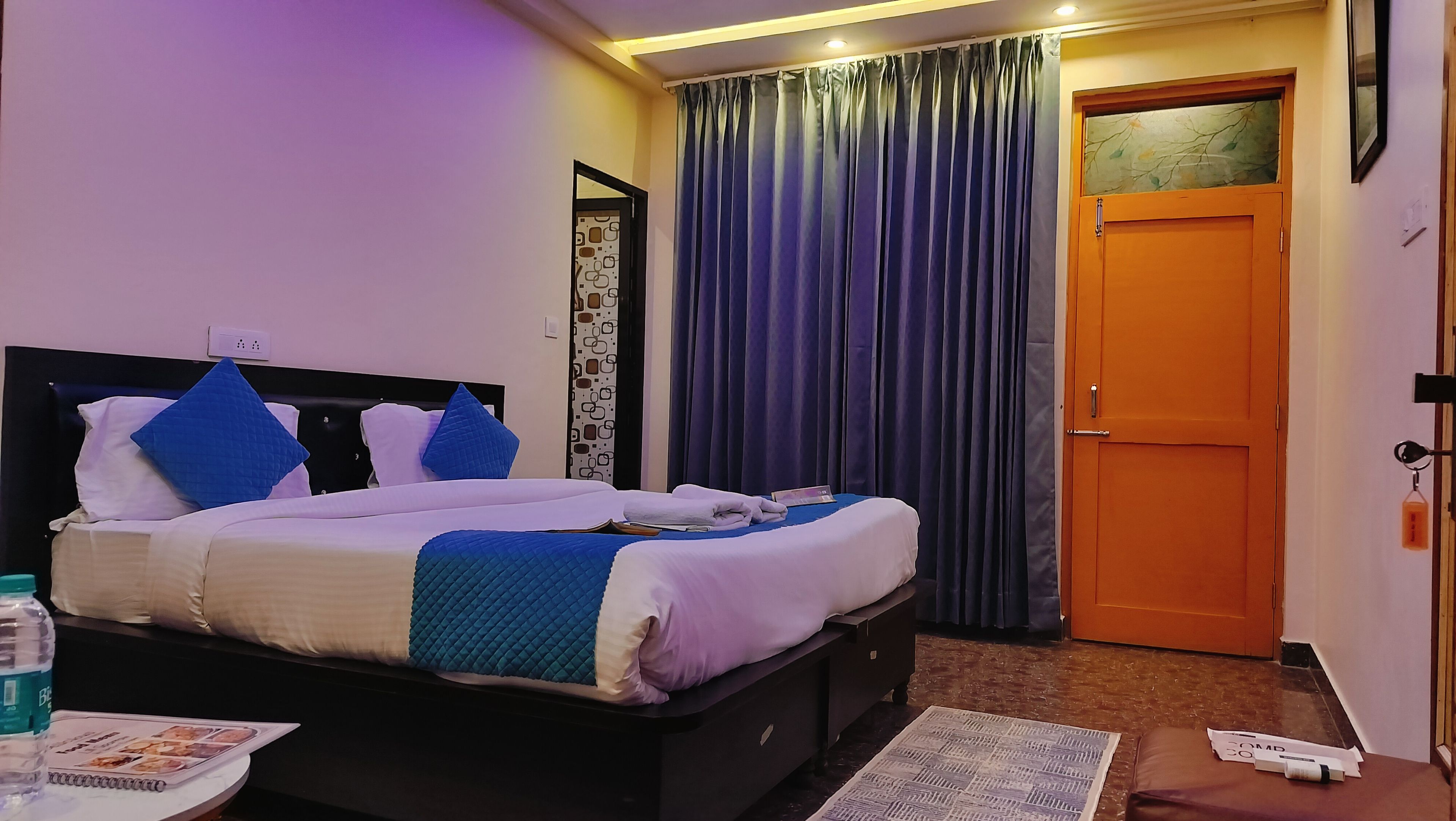 Una habitación de hotel con cama, mesita de noche, puerta y ventana con cortinas.