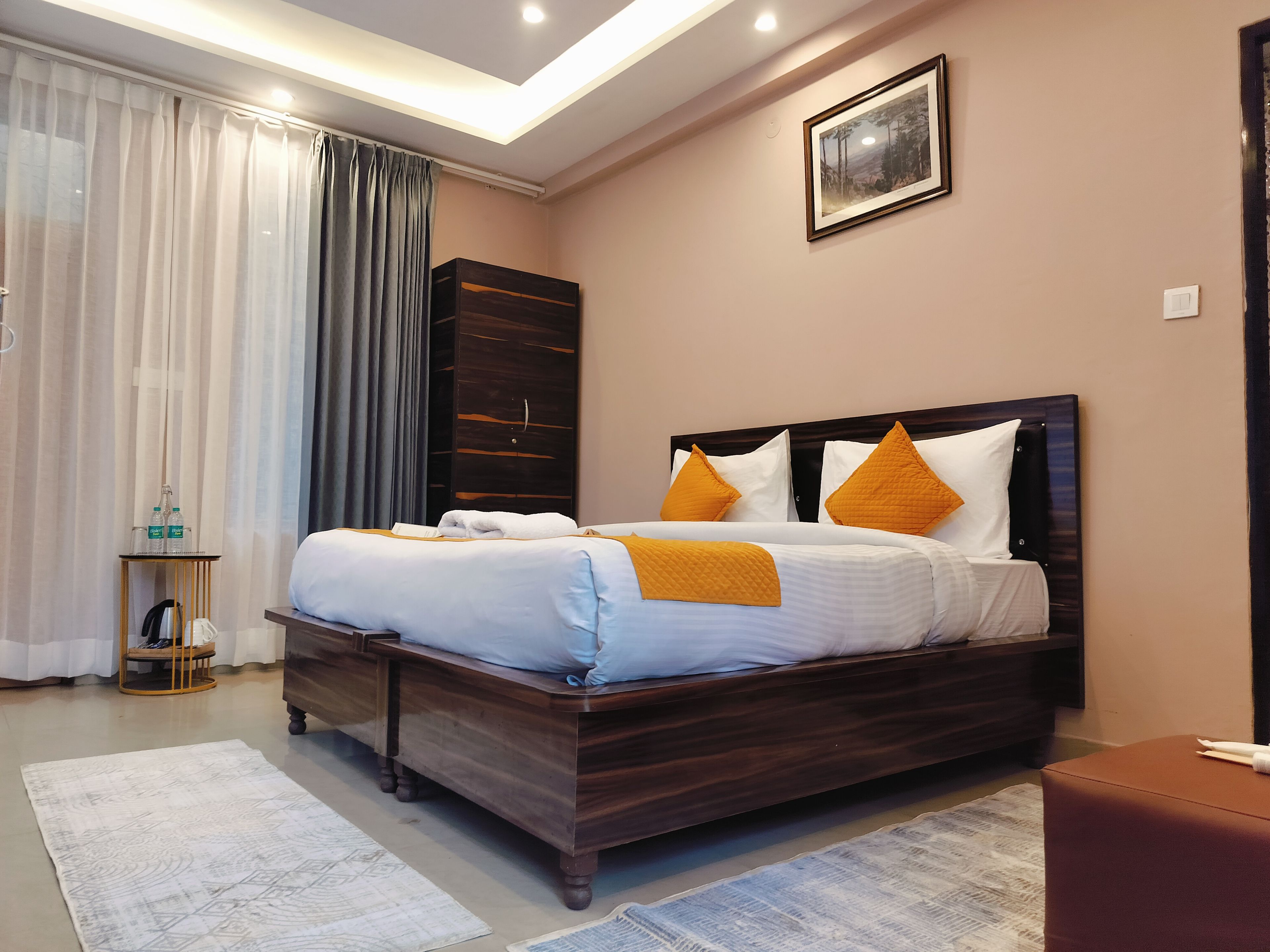 A neatly made bed with a wooden headboard, a bedside table with a bottle and a cup, and a large window with curtains.