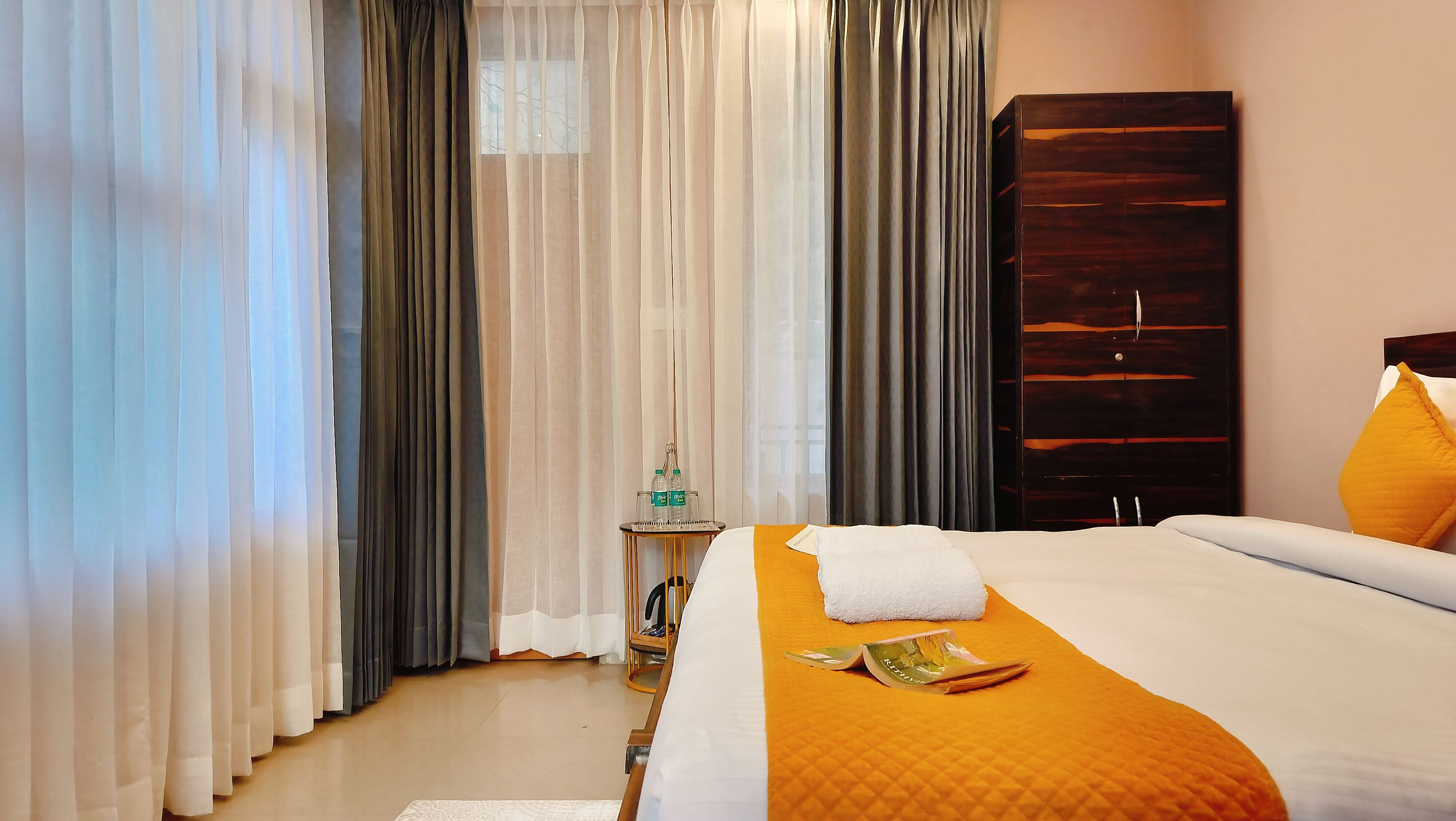 A hotel room with a bed, a wooden wardrobe, and a small table with bottles and glasses.