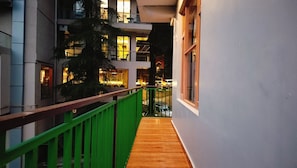 Un pasillo con piso de madera, barandilla verde y vistas a un edificio con múltiples ventanas y luces.