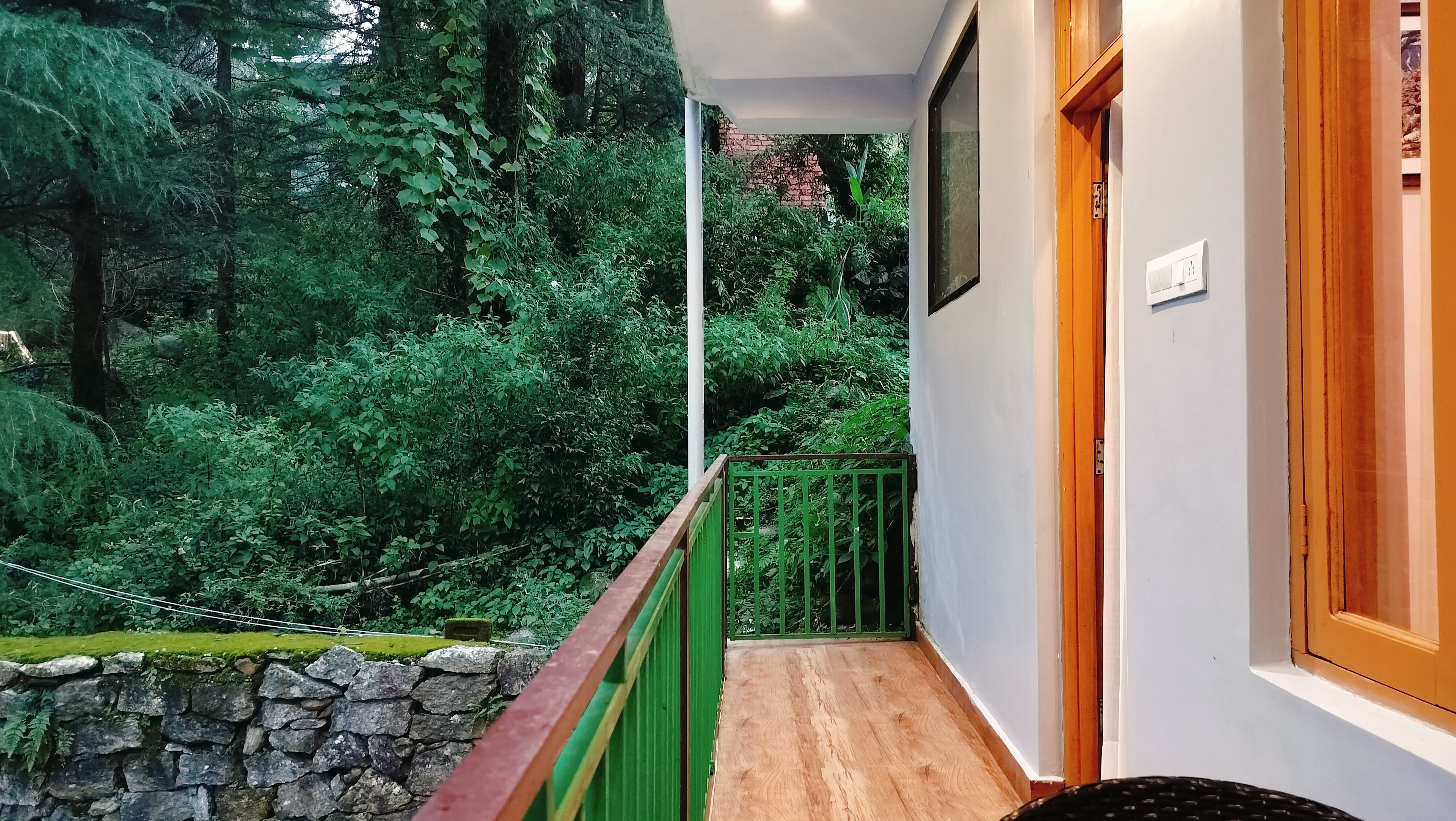 Un balcón con pared de piedra, barandilla verde y vista a abundante vegetación.