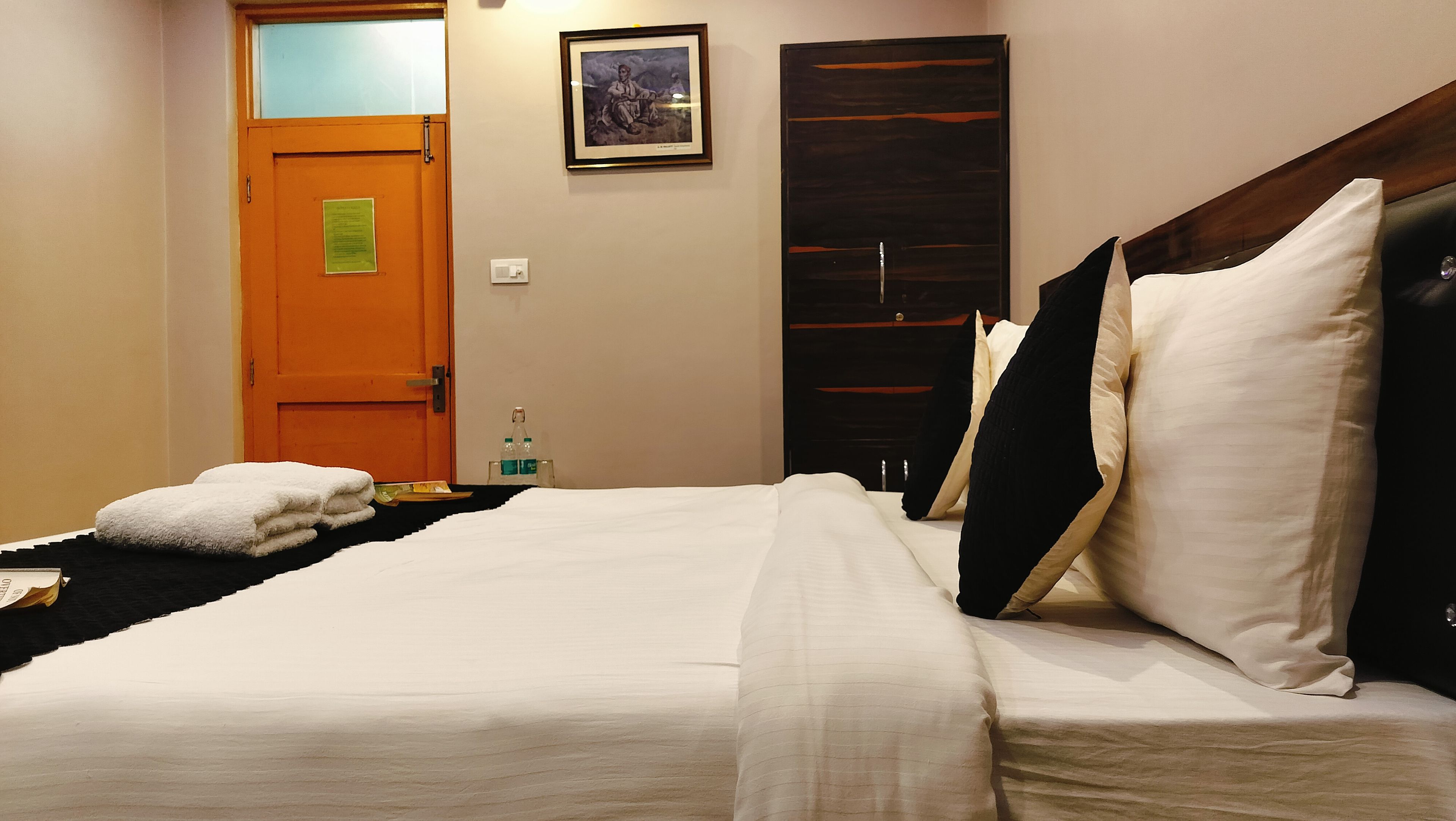 A hotel room with a bed, a door, a framed picture on the wall, and a small table with a bottle.