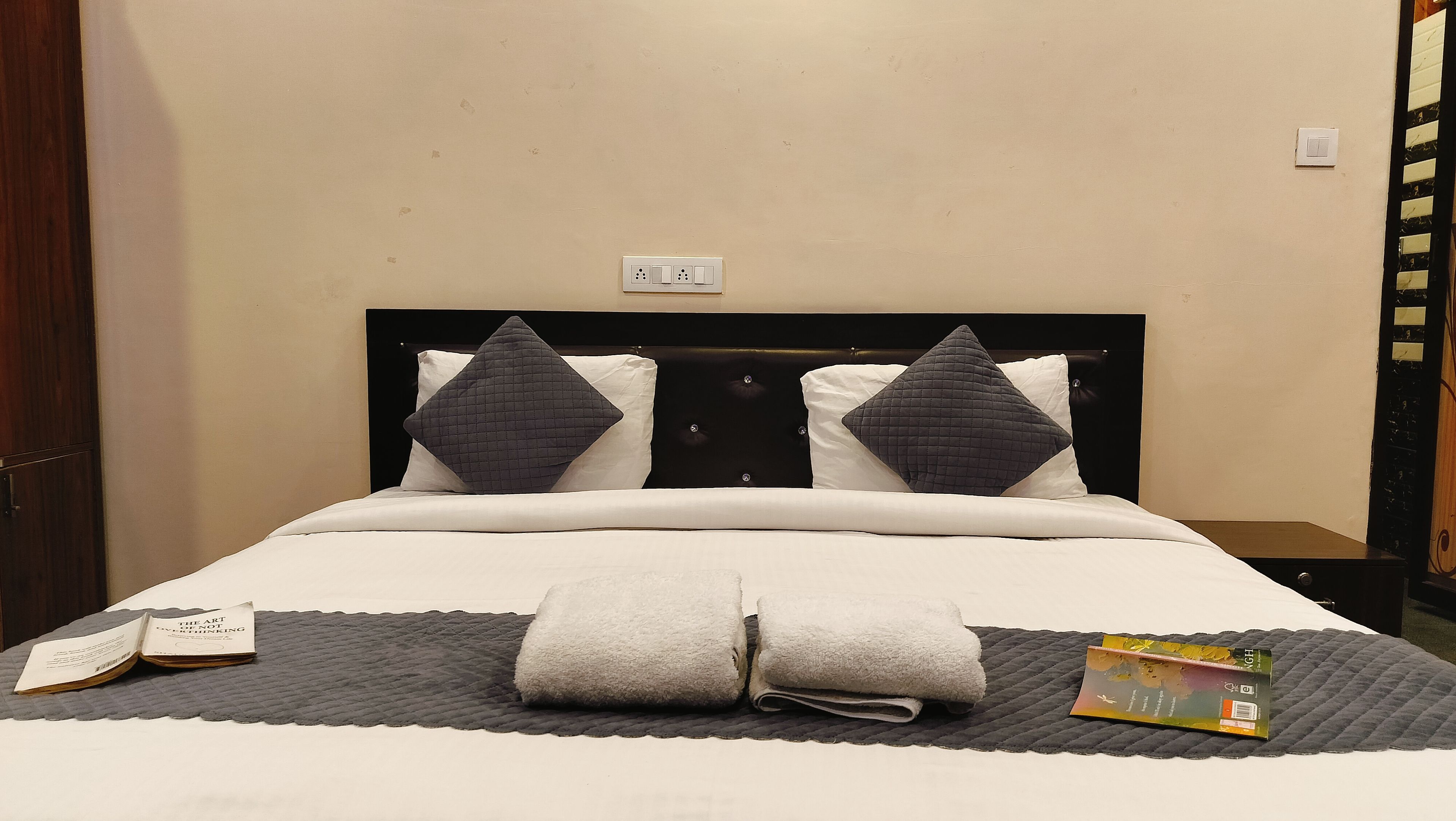 A hotel room with a bed, pillows, a bedside table, a book, and a power outlet.