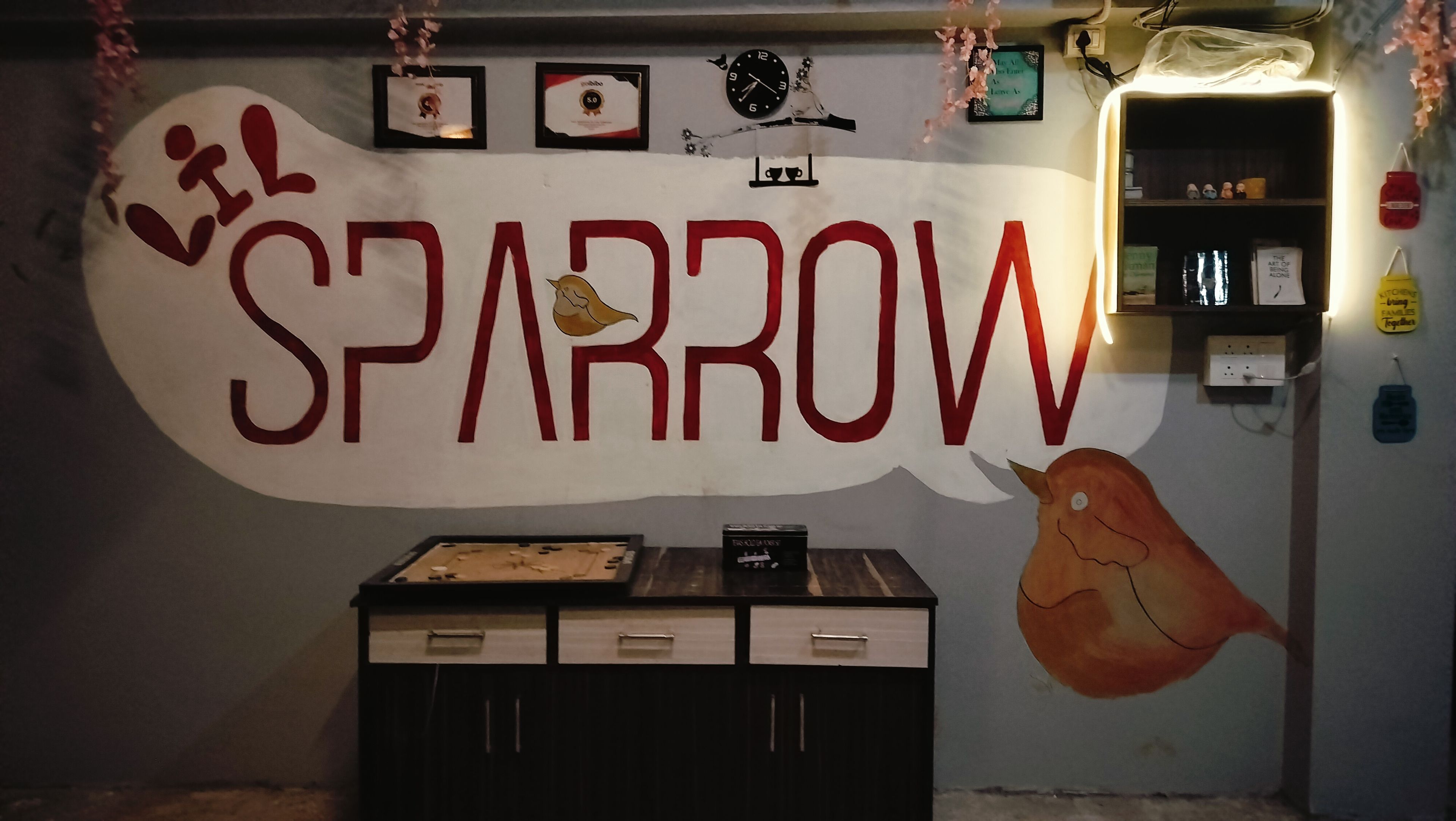 Una pared con la palabra 'SPARROW' y una ilustración de un pájaro, un reloj, certificados enmarcados y una estantería con objetos.