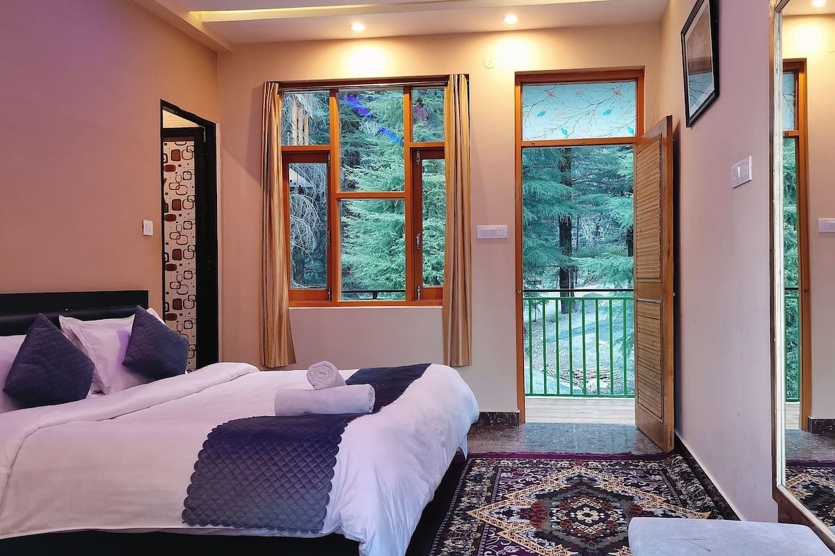 Habitación de hotel con cama, un pequeño rincón de descanso, balcón con vistas a árboles y puerta que da al exterior.