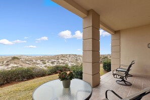 Un patio con una mesa de vidrio y dos sillas, con vista a una playa con dunas y cielo despejado.