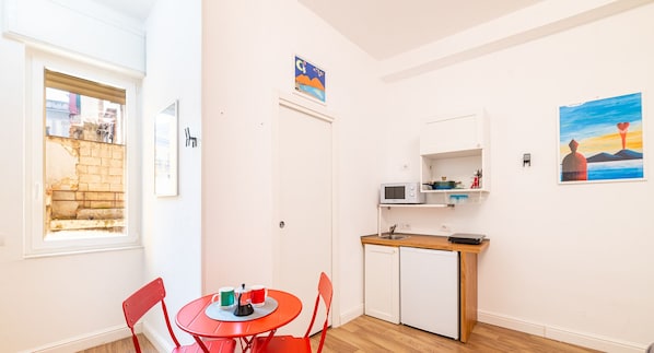 A compact kitchen with a microwave, sink, and cabinets, a small dining table with red chairs, and a window with a view of a brick wall. - Tarantella By DomusExtra (Naples)