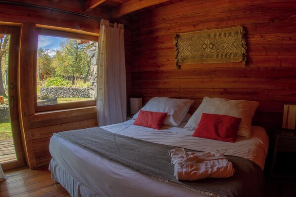 A wooden cabin bedroom with a bed, pillows, and a view of the outdoors. - Rocanegra Mountain Lodge (Pinto)
