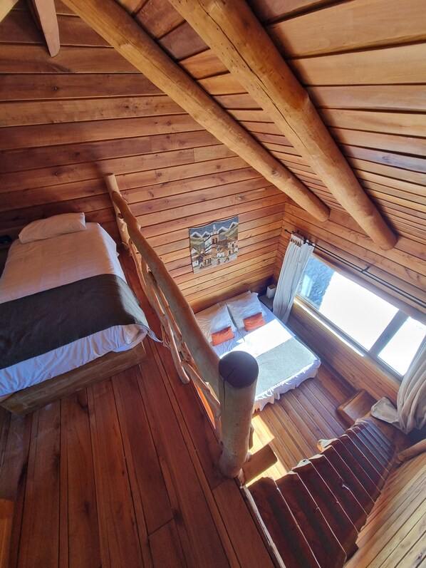 A wooden cabin with a loft bedroom featuring two beds, a window, and a wooden ceiling.