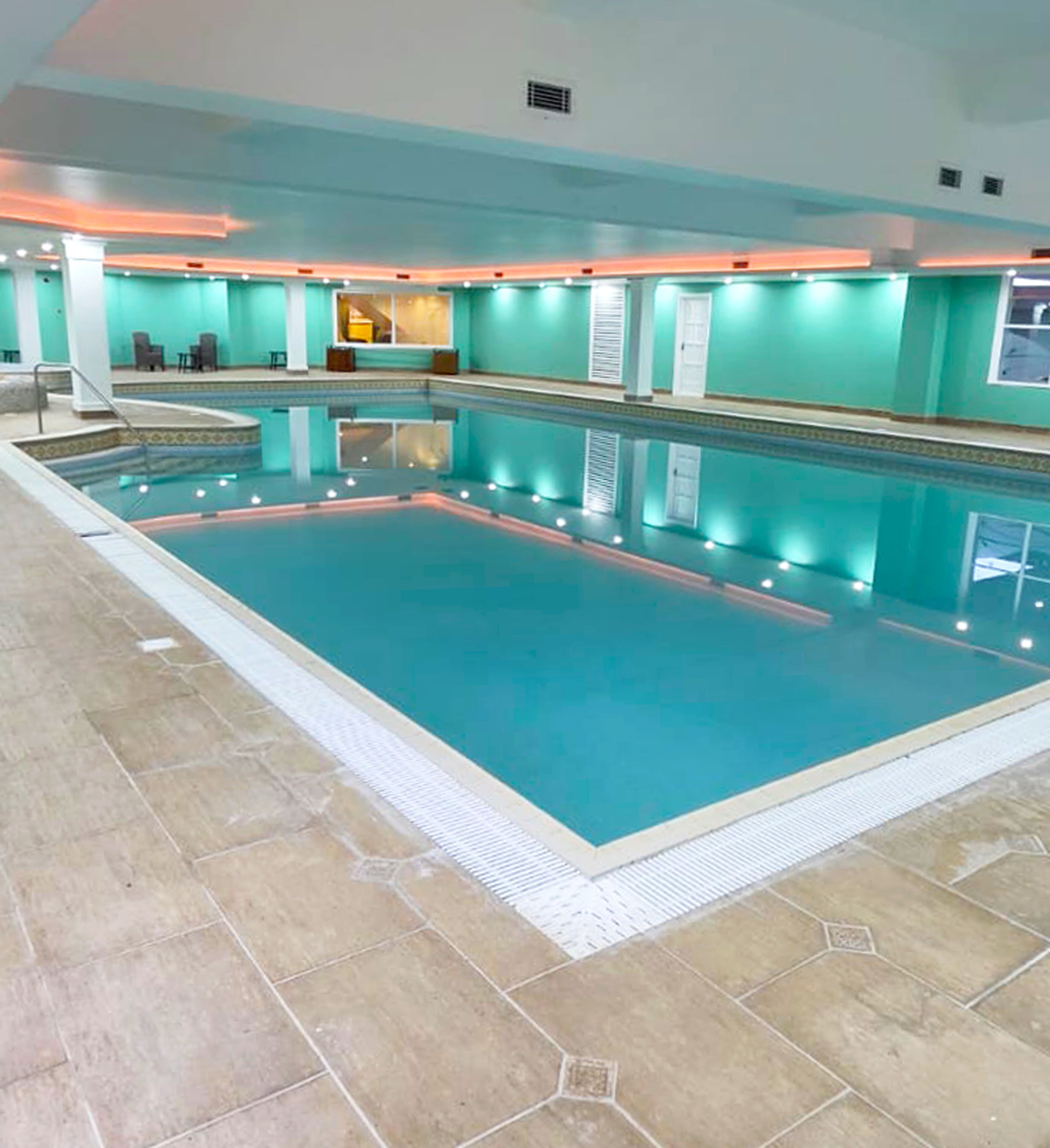 An indoor swimming pool with clear water, surrounded by a tiled floor and walls, and a seating area with chairs and tables.