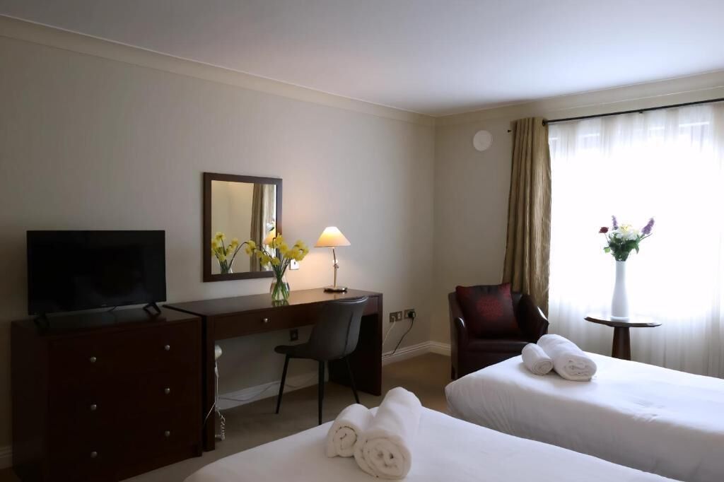 A hotel room with two beds, a desk with a chair, a television, a lamp, and a vase with flowers.