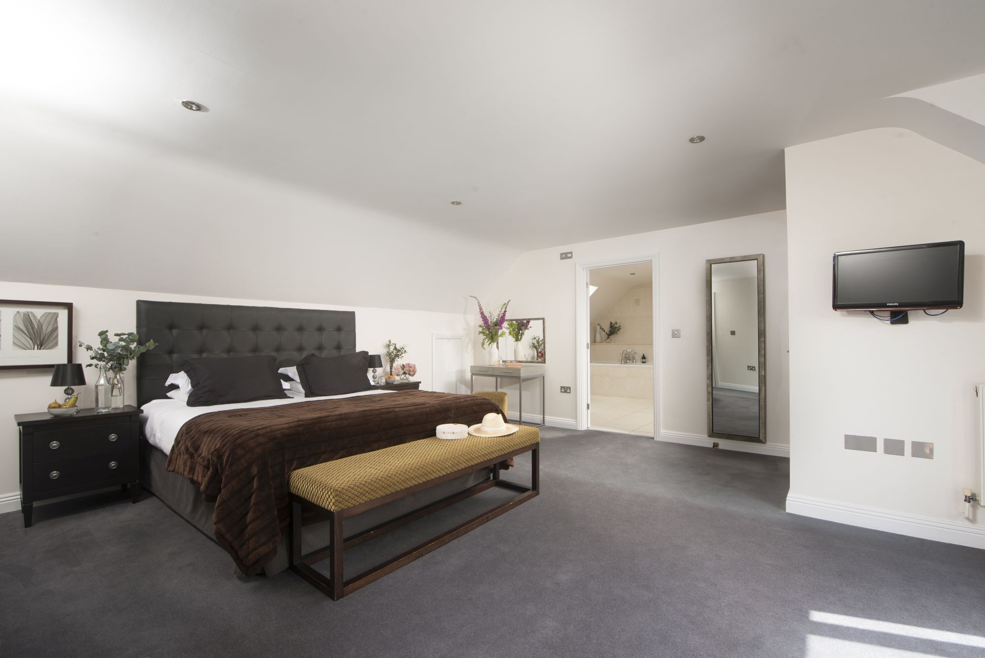 Une chambre à coucher moderne avec un grand lit, des tables de chevet, un banc, un miroir et un téléviseur.
