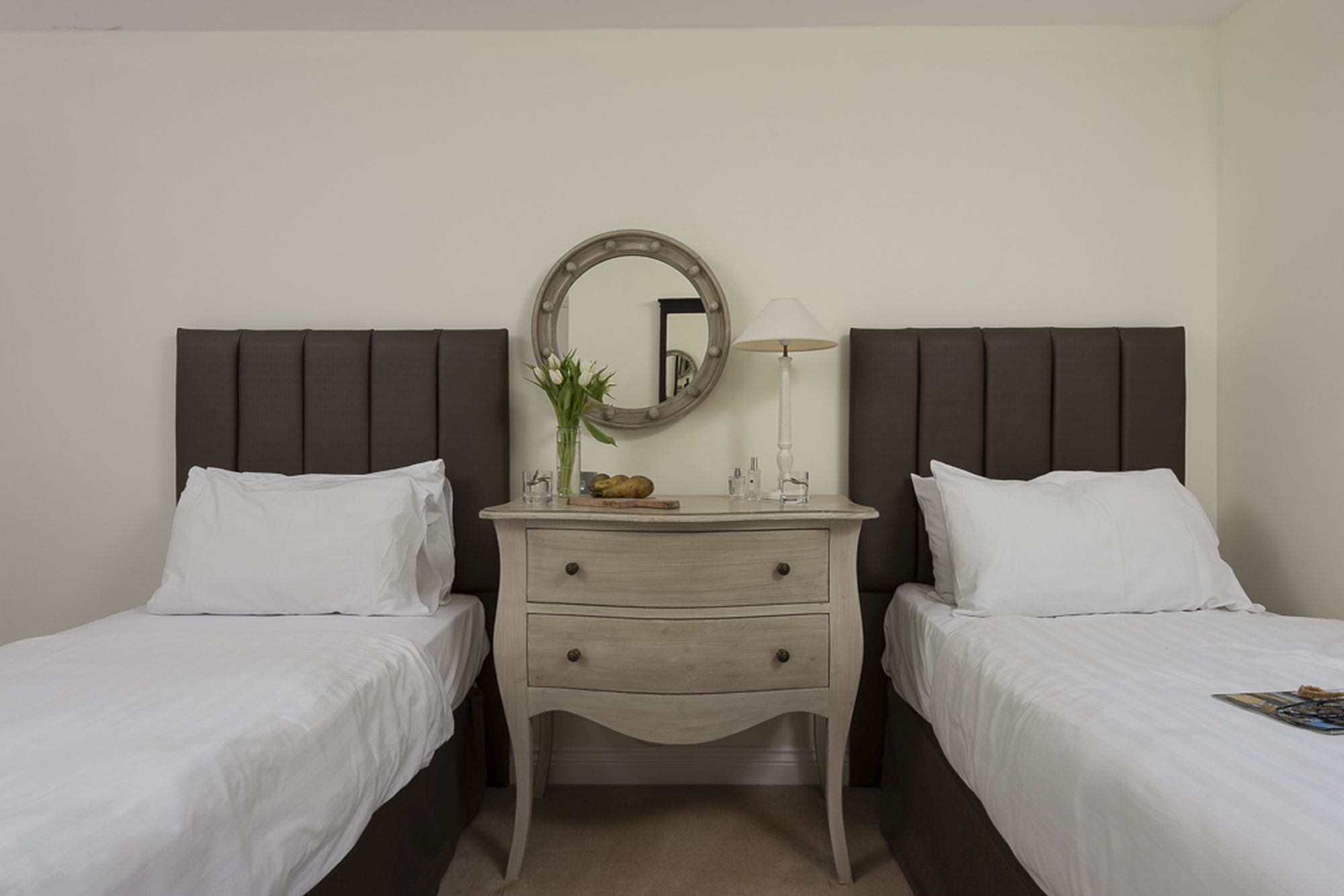 A hotel room with two single beds, a bedside table with a lamp, a mirror, and a small vase with flowers.