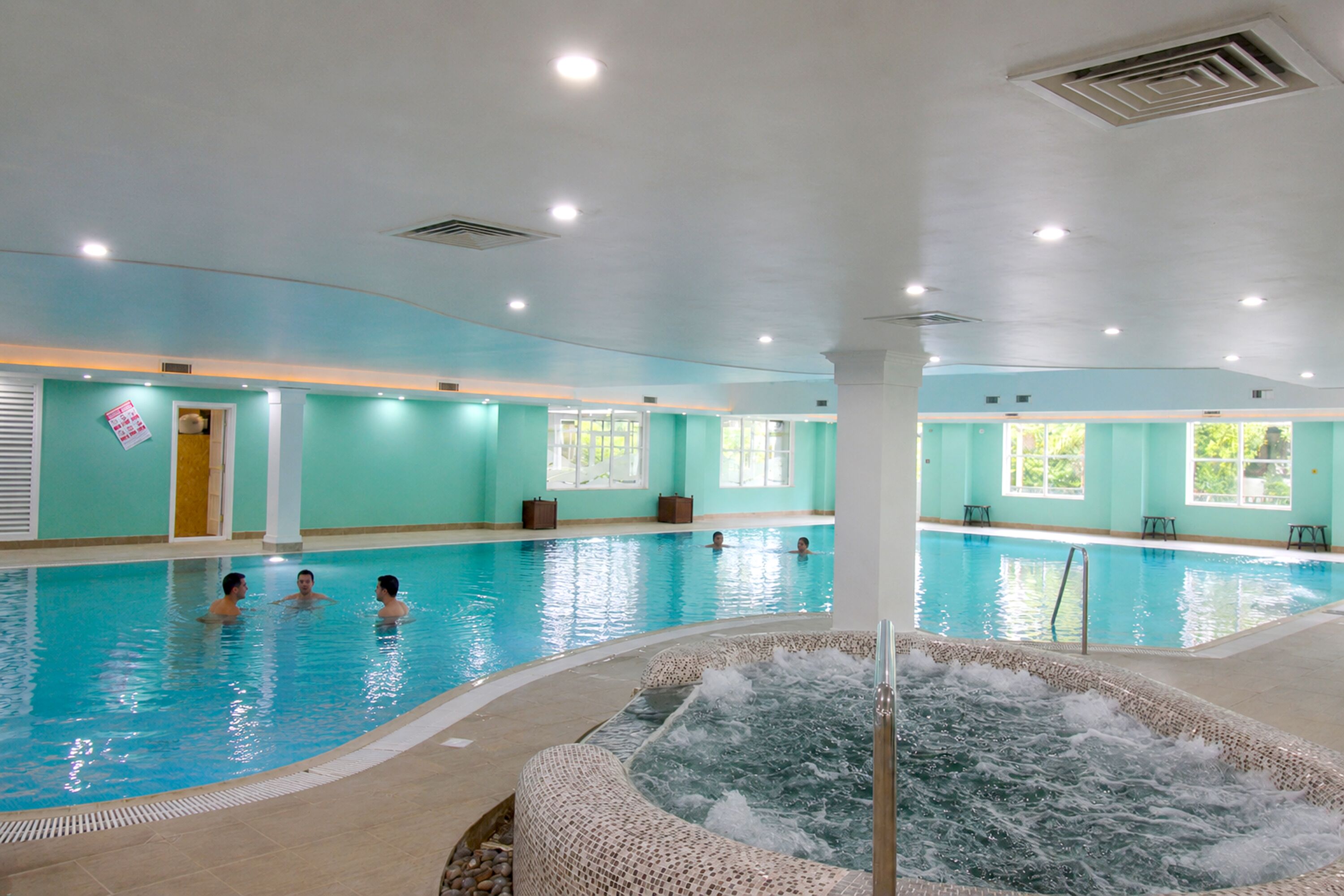 An indoor swimming pool with a hot tub, tiled walls, and ceiling lights.