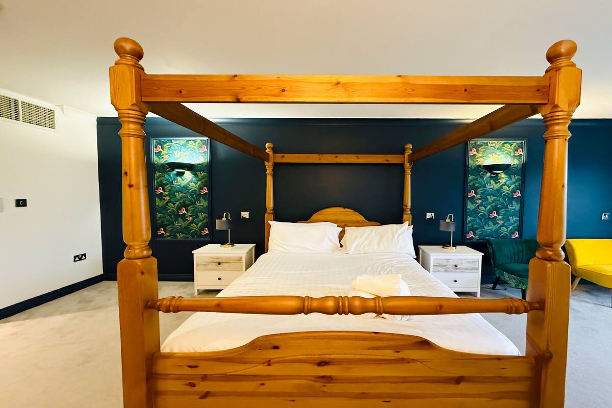 A four-poster bed with a wooden frame and white bedding, flanked by nightstands with lamps, in a room with dark walls and floral wallpaper.