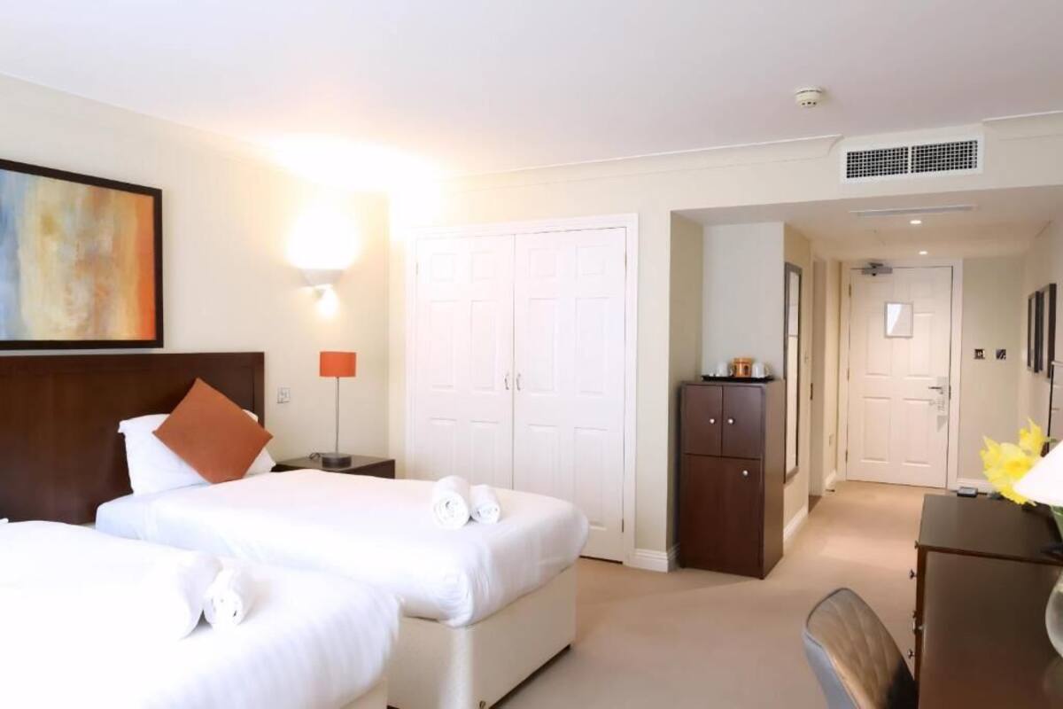 A hotel room with two beds, a desk, a wardrobe, and a painting on the wall.