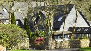 A stone-walled building with a sloped roof, surrounded by greenery and a paved driveway.