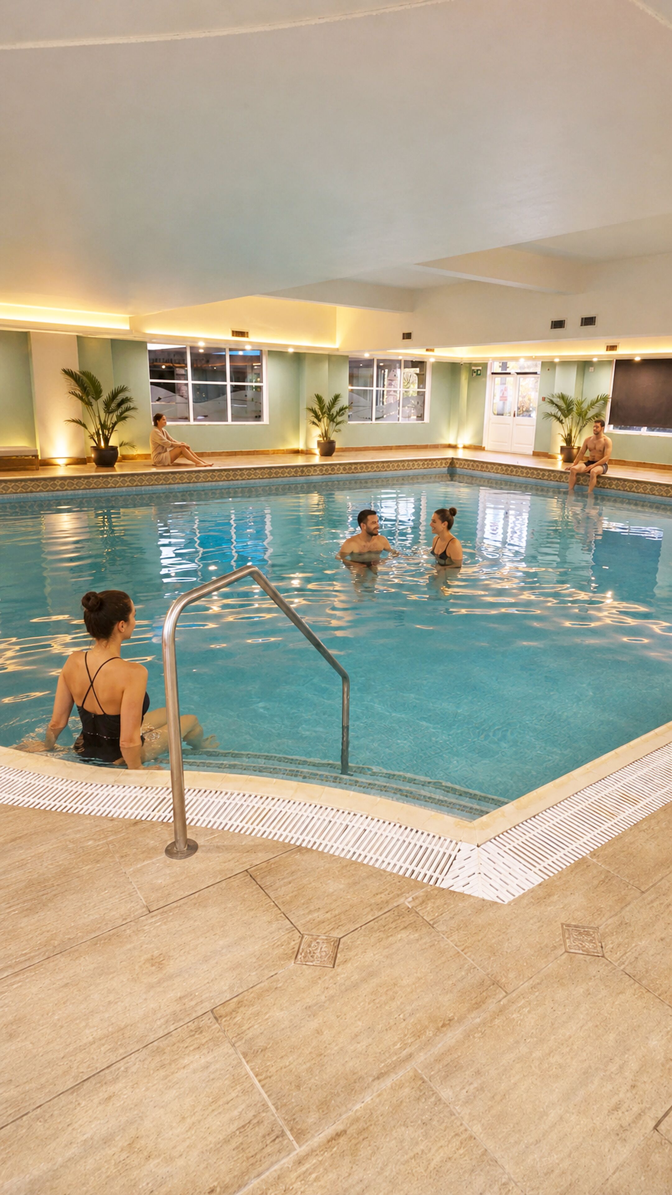 An indoor swimming pool with clear water, a tiled floor, and potted plants around the perimeter.