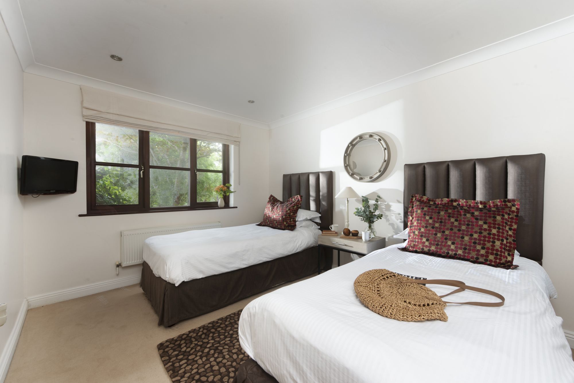 Quarto de hotel com duas camas, uma televisão, uma janela, um espelho e uma mesa de cabeceira com abajur e um vaso de flores.