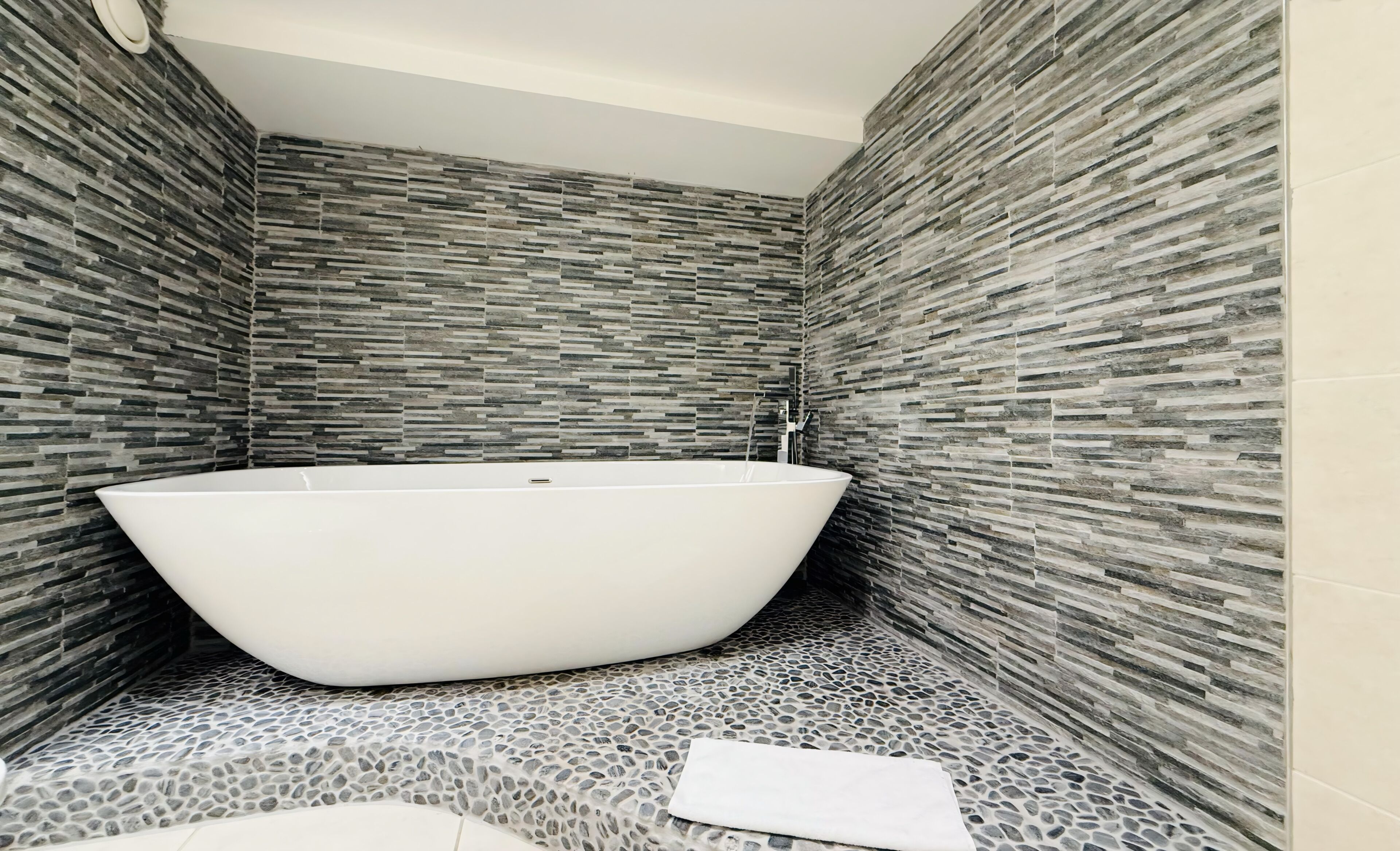 A modern bathroom with a freestanding bathtub, stone mosaic flooring, and textured wall tiles.
