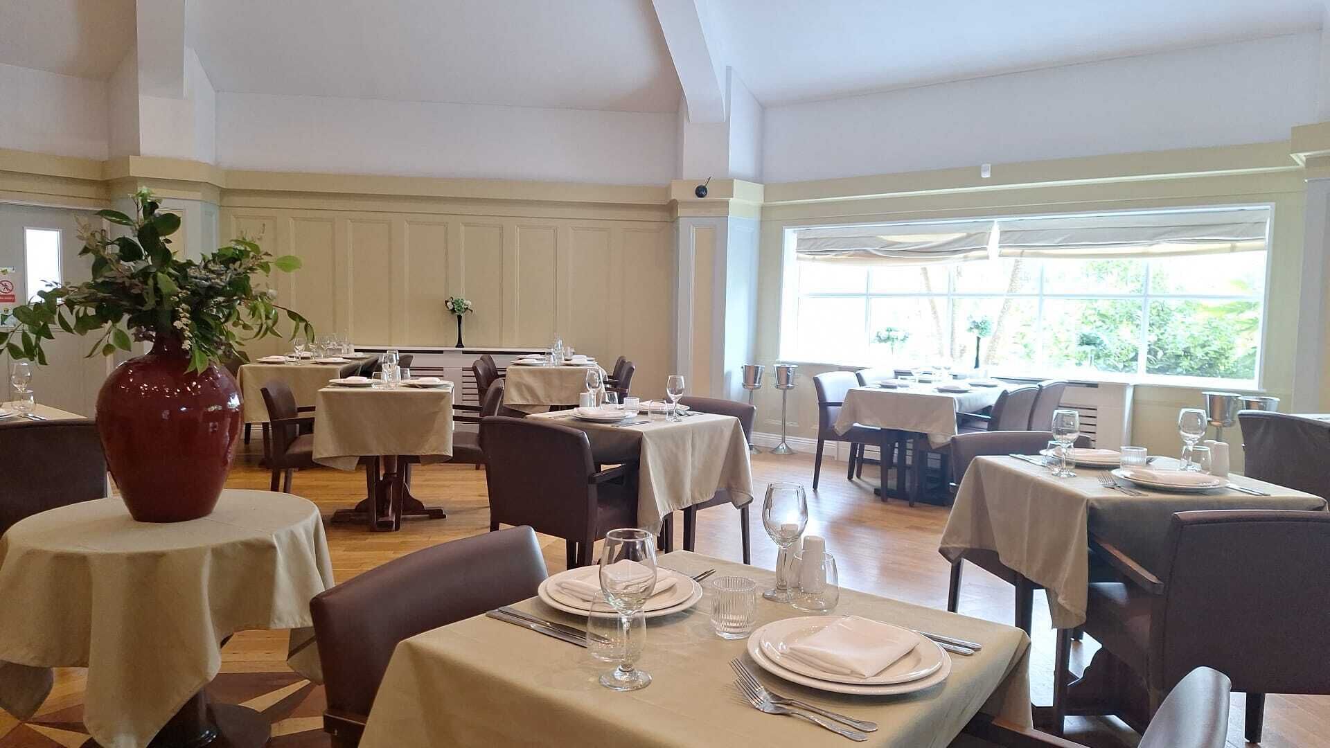A dining area with multiple tables set for service, a large vase with flowers, and a view of greenery through the windows.