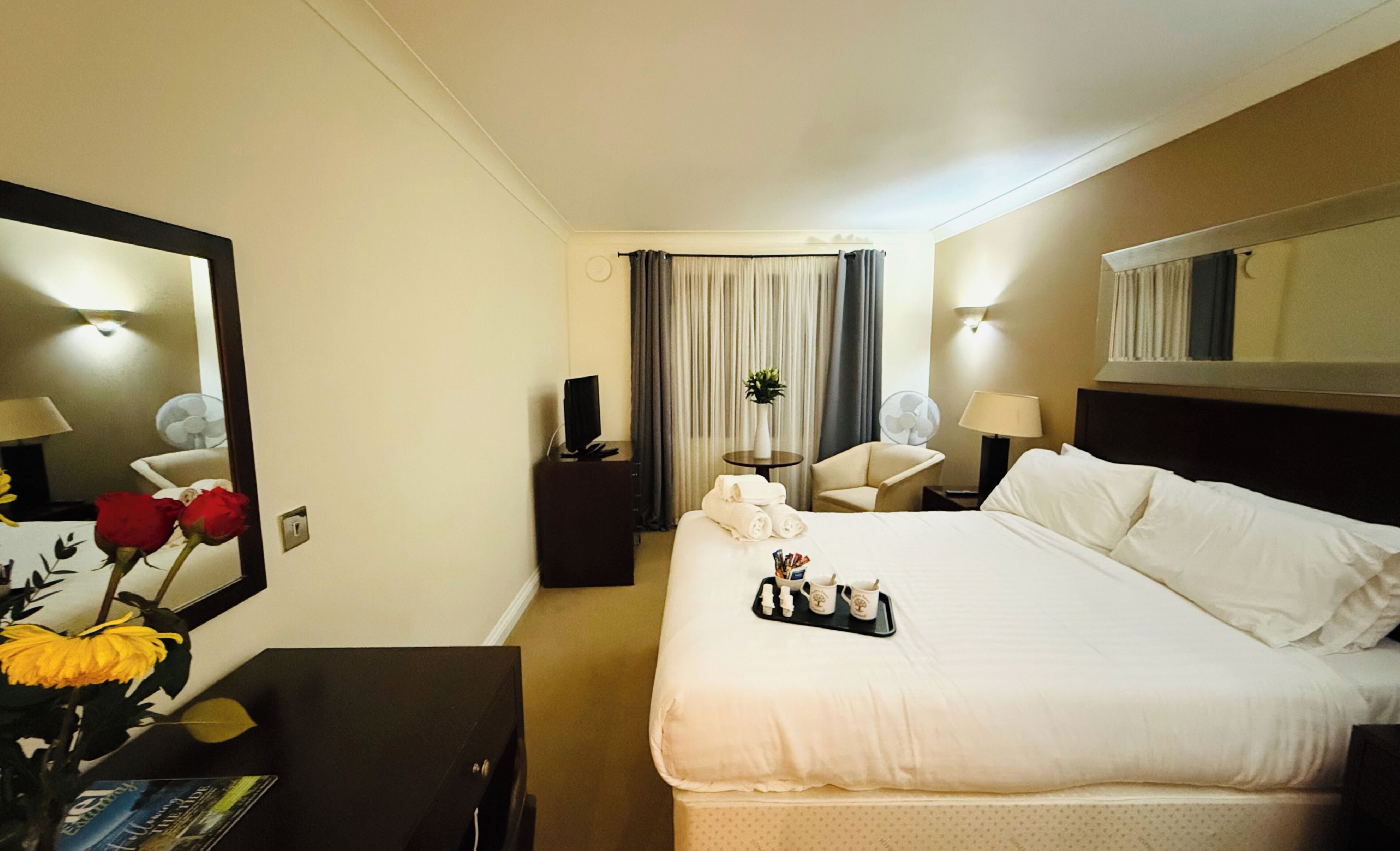 Een hotelkamer met een groot bed, een bureau met een lamp, een stoel, een spiegel en een vaas met bloemen.