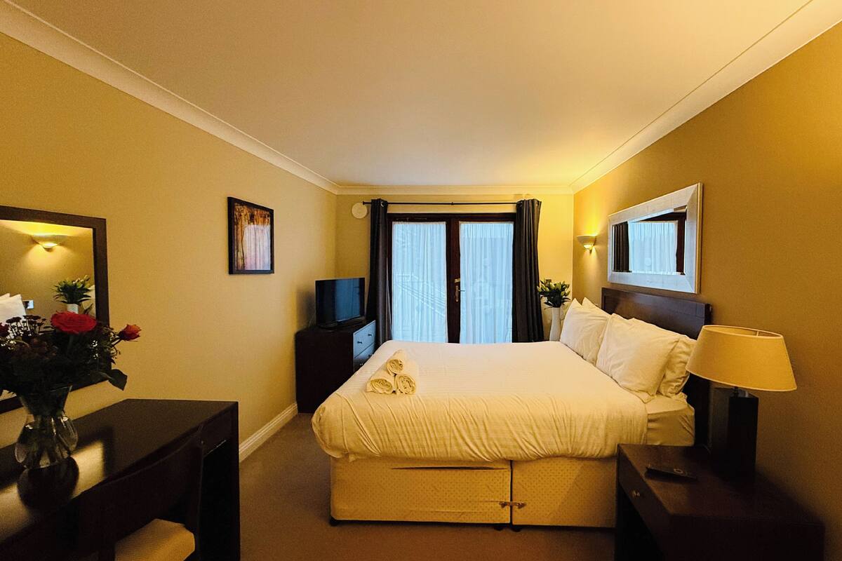 A hotel room with a bed, a bedside table, a lamp, a TV, a mirror, and a window with curtains.