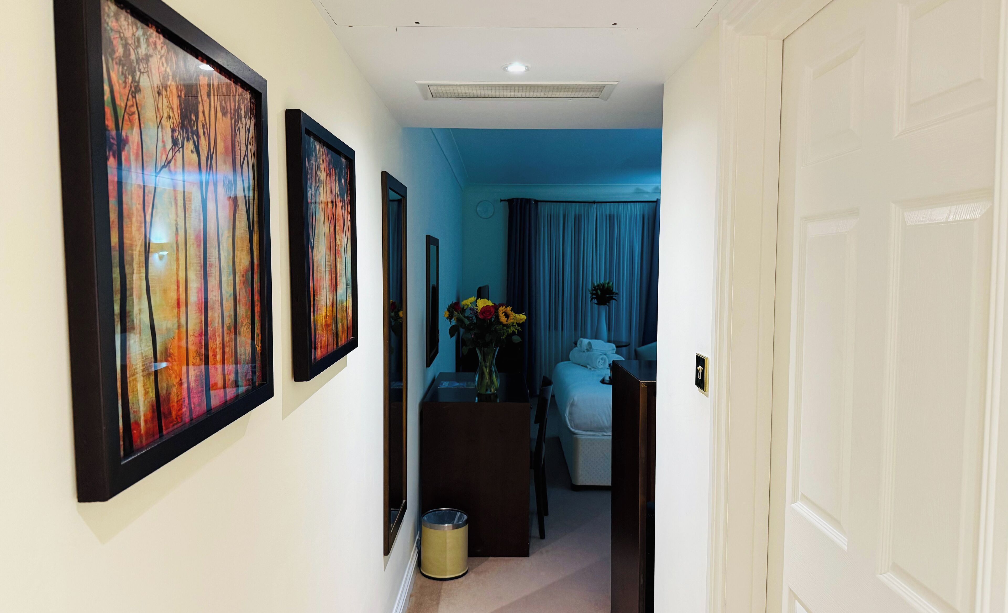 A hallway with framed artwork on the walls, a desk with a vase of flowers, and a bed with a blue curtain in the background.
