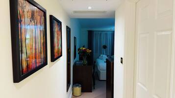 A hallway with framed artwork on the walls, a desk with a vase of flowers, and a bed with a blue curtain in the background.