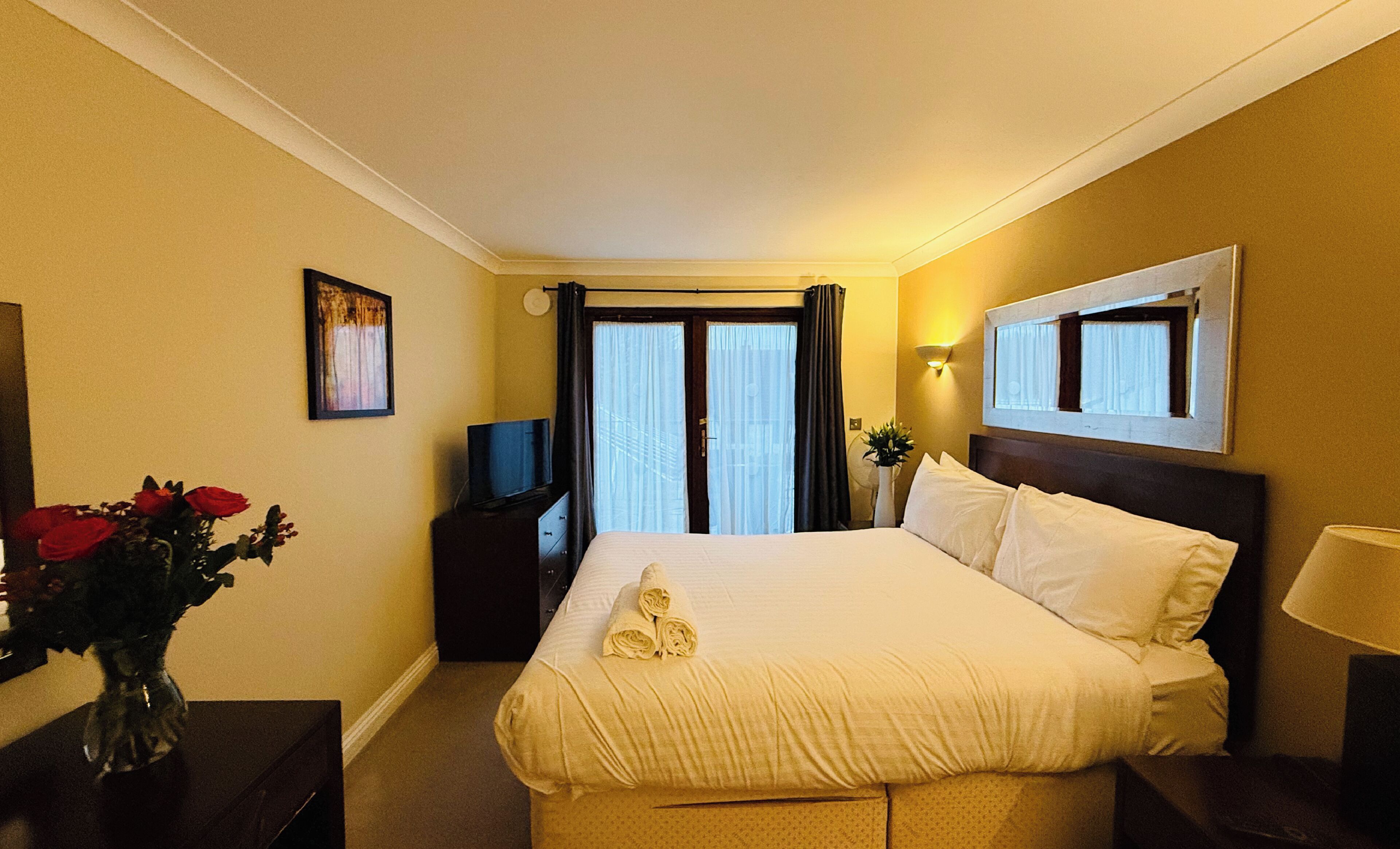 Una habitación de hotel con una cama, un televisor, un jarrón con flores rojas y un ventanal con cortinas.
