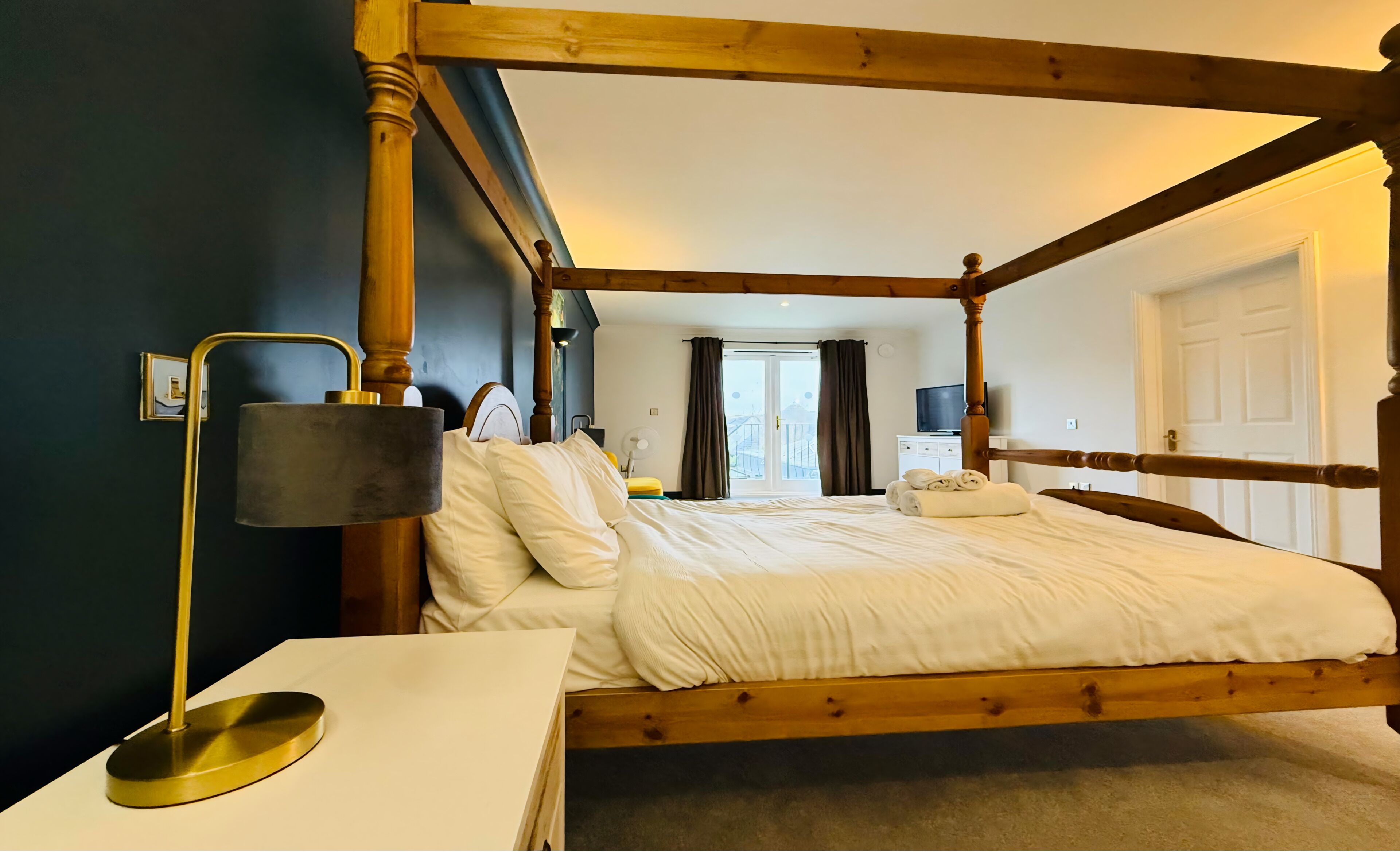 A four-poster bed with a wooden frame, a bedside table with a lamp, and a view of the outside through a window.