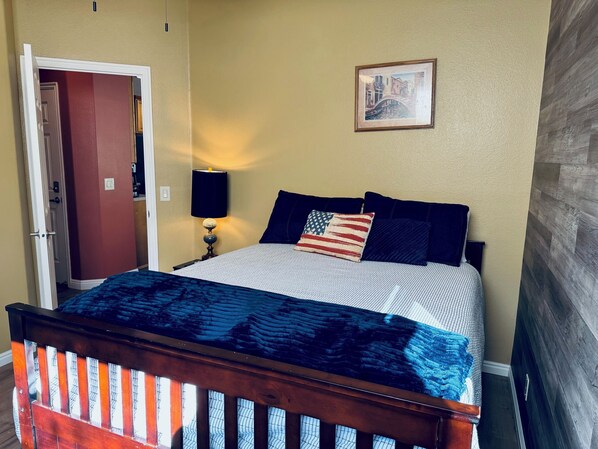A bedroom with a bed, a nightstand, a lamp, a framed picture on the wall, and a door to another room.
