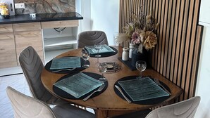 A dining area with a wooden table set for four, surrounded by upholstered chairs. A kitchen with wooden cabinets and a marble backsplash is visible in the background. - Le Corossol (Fort-de-France)