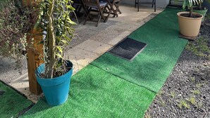 A covered patio area with a table and chairs, surrounded by potted plants and a green artificial turf path. - Le Corossol (Fort-de-France)