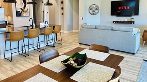 Una sala de estar moderna con un sofá gris, un comedor con una mesa y sillas de madera, una cocina con luces colgantes y un televisor que muestra 'NETFLIX'.