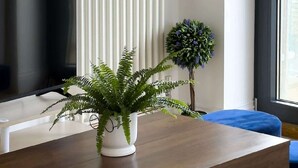 Un salon moderne avec un téléviseur à écran plat, un radiateur blanc, une table basse en bois, une fougère en pot et un coussin bleu.
