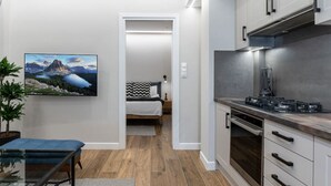 Una cuina moderna amb electrodomèstics d'acer inoxidable, una zona de menjador amb taula de vidre i un dormitori amb llit i tauleta de nit.