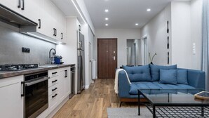 Una cuina moderna amb armaris blancs, un forn de dipòsit i una campana extractora d'acer inoxidable. A la zona de sala hi ha un sofà blau i una taula de centre de vidre.