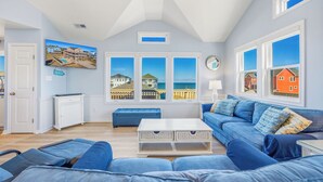 Een woonkamer met een groot raam, een blauwe bank, een witte salontafel en een televisie aan de muur.