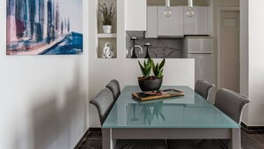 A modern dining area with a glass table, grey upholstered chairs, a potted plant, and a cityscape painting on the wall. - Beachfront apartment in Voula w/balcony (Voula)