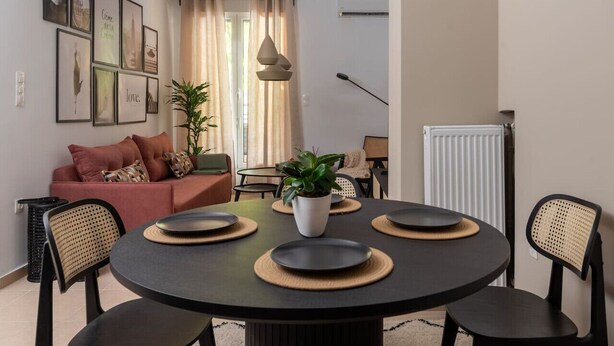 Una sala de estar moderna con una mesa de comedor redonda, sillas negras y un sofá con cojines. Se observan cuadros enmarcados en la pared, una planta en maceta y una ventana con cortinas.