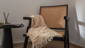 Una silla con respaldo tejido y una manta beige, una mesita redonda con base negra, y un gato acostado en el suelo.