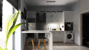 Een moderne keuken met een wit aanrecht, zwarte barkrukken, een ingebouwde wasmachine en een groot raam met een plant.