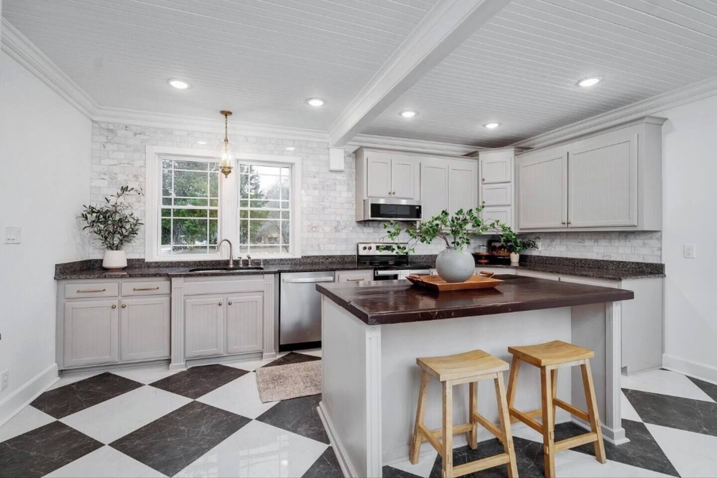 Une cuisine avec des meubles blancs, un îlot central doté de tabourets, un carrelage noir et blanc à motifs et une fenêtre donnant sur un espace verdoyant.