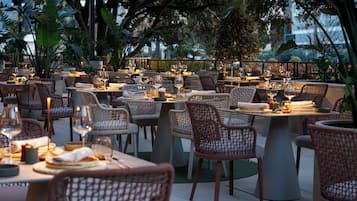 An outdoor dining area with wicker chairs and tables, surrounded by greenery.